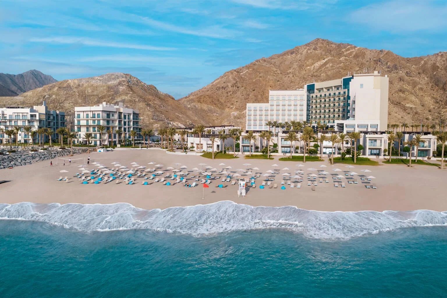 A beachfront resort with rows of sun loungers and umbrellas on a sandy beach, set against a backdrop of rocky hills and a clear blue sky.
