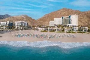 A beachfront resort with rows of sun loungers and umbrellas on a sandy beach, set against a backdrop of rocky hills and a clear blue sky.