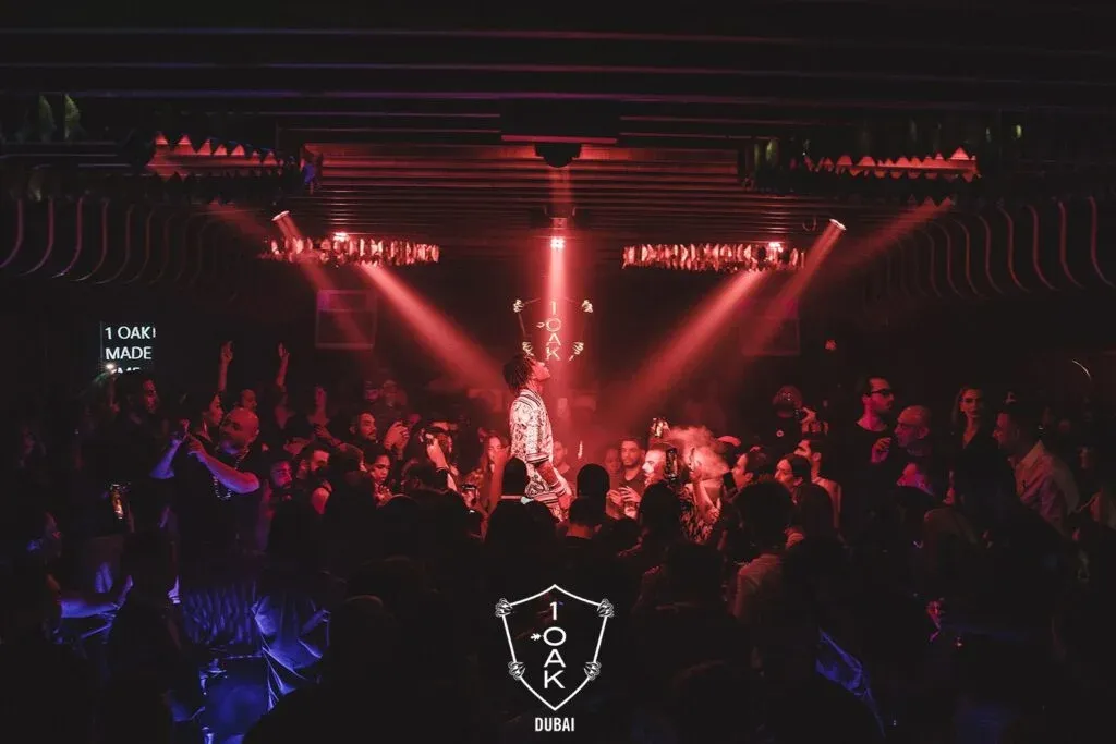 A crowded nightclub scene with red lighting, featuring a performer on stage at 1OAK Dubai. The audience is lively, with many holding up phones to capture the moment.