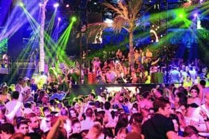 Crowded outdoor nightclub scene with people dancing under colorful stage lights and palm trees.