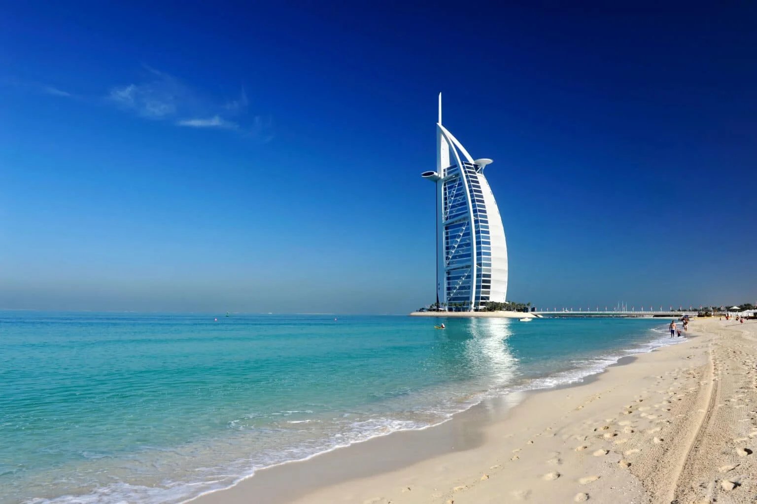 A modern sail-shaped skyscraper near a sandy beach with turquoise water under a clear blue sky.