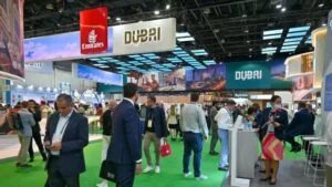 People walking and interacting at a trade show with stands displaying "Dubai" and "Emirates" signs, featuring various promotional materials and a bright ceiling display.