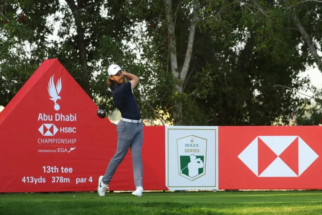 A golfer in mid-swing on the 13th tee at the Abu Dhabi HSBC Championship, surrounded by red signage with tournament branding and sponsorship logos, amidst a backdrop of trees.