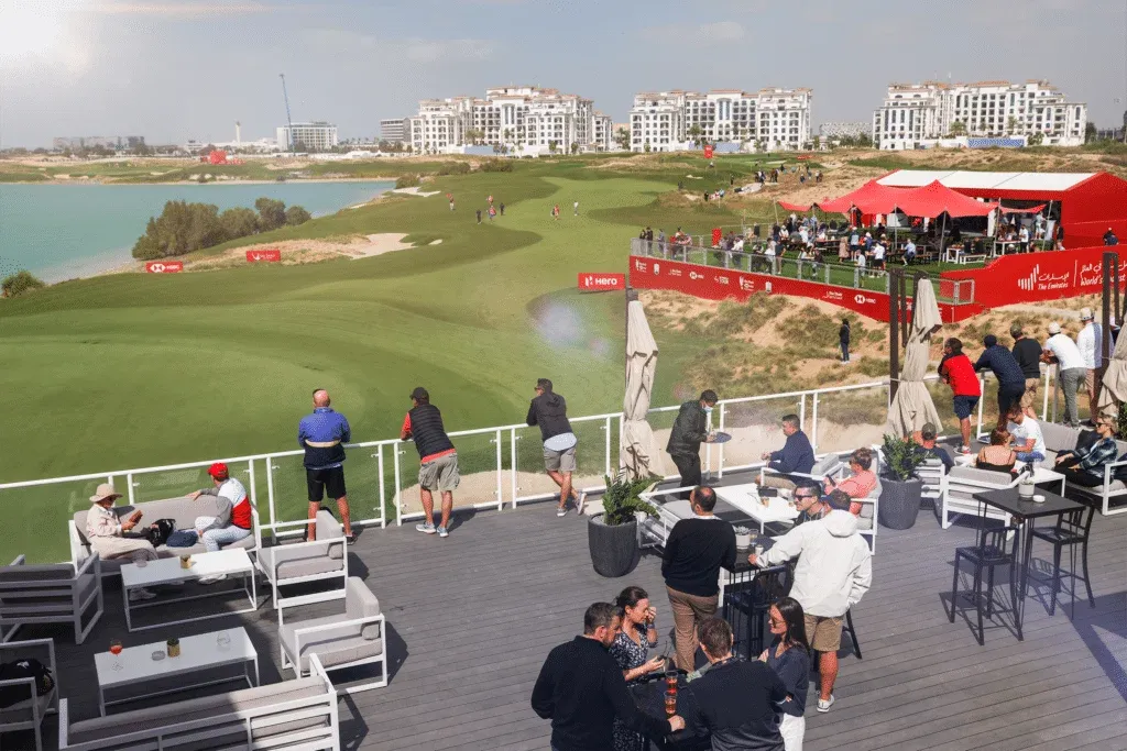 A stylish outdoor viewing area overlooking a golf course with a large grass field, surrounded by spectators and buildings in the background. People are sitting and standing on a deck, enjoying drinks and conversation, while watching the golf event. A red hospitality enclosure is visible to the right with more people observing the game.