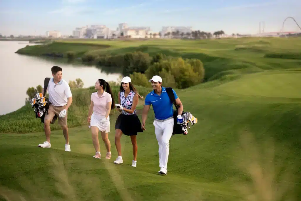 Four people walking on a golf course, carrying golf bags, with a lake and cityscape in the background.