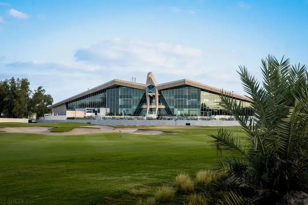 Modern building with large glass windows and triangular roof, set against a blue sky, with a grassy field and palm trees in the foreground.