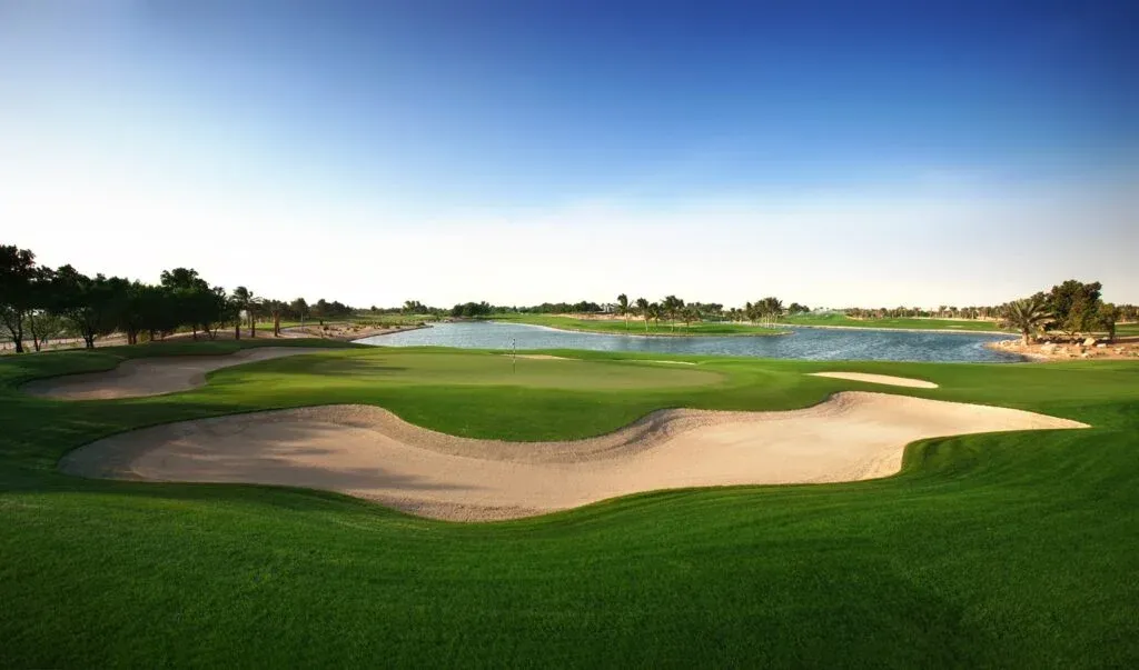 A lush green golf course with a sand bunker in the foreground and a water hazard further back, surrounded by trees under a clear blue sky.