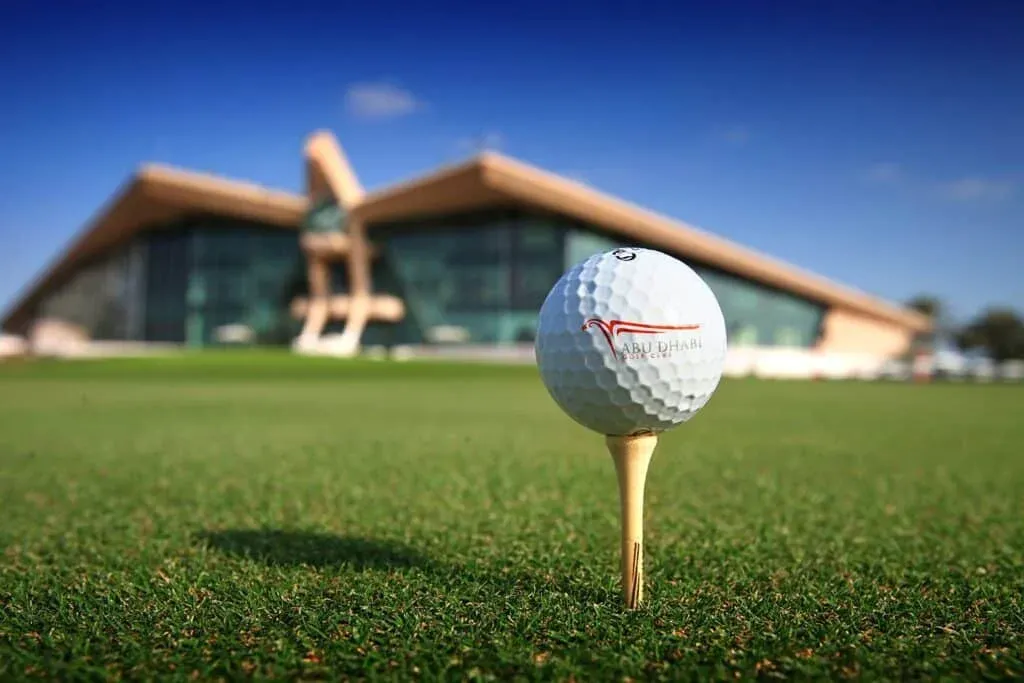 Close-up of a golf ball with the Abu Dhabi Golf Club logo on a tee, set against a blurred background of a building with a distinctive wing-shaped roof.