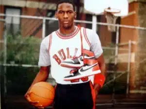 A person wearing a Chicago Bulls jersey holding a basketball and a pair of red and black Nike sneakers over their shoulder, standing on an outdoor basketball court.