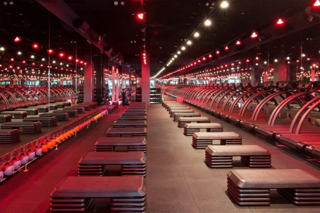 A dimly lit gym with rows of treadmills on one side and stacked benches on the other, illuminated by red lights, with walls lined with mirrors.