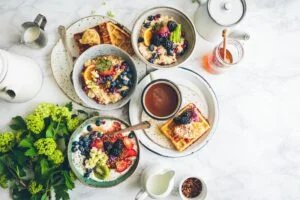 A breakfast spread featuring bowls of oatmeal topped with assorted fruits like blueberries, blackberries, strawberries, kiwi, and flowers, accompanied by waffles with honey and fruit, and a cup of coffee, set on a marble table with jars of milk and honey, and green floral arrangements.