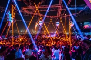 A crowded nightclub with vibrant orange and blue stage lights illuminating the space, set against a night city skyline in the background.