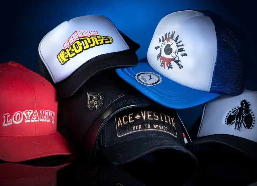 A collection of six caps with various designs, including a red cap with the word 'Loyalty,' a black cap with an embroidered logo, a white and black cap featuring 'My Hero Academia' text in Japanese, a blue and white cap with 'Adrenaline Freak' text, and caps with other unique designs, all set against a dark blue background.