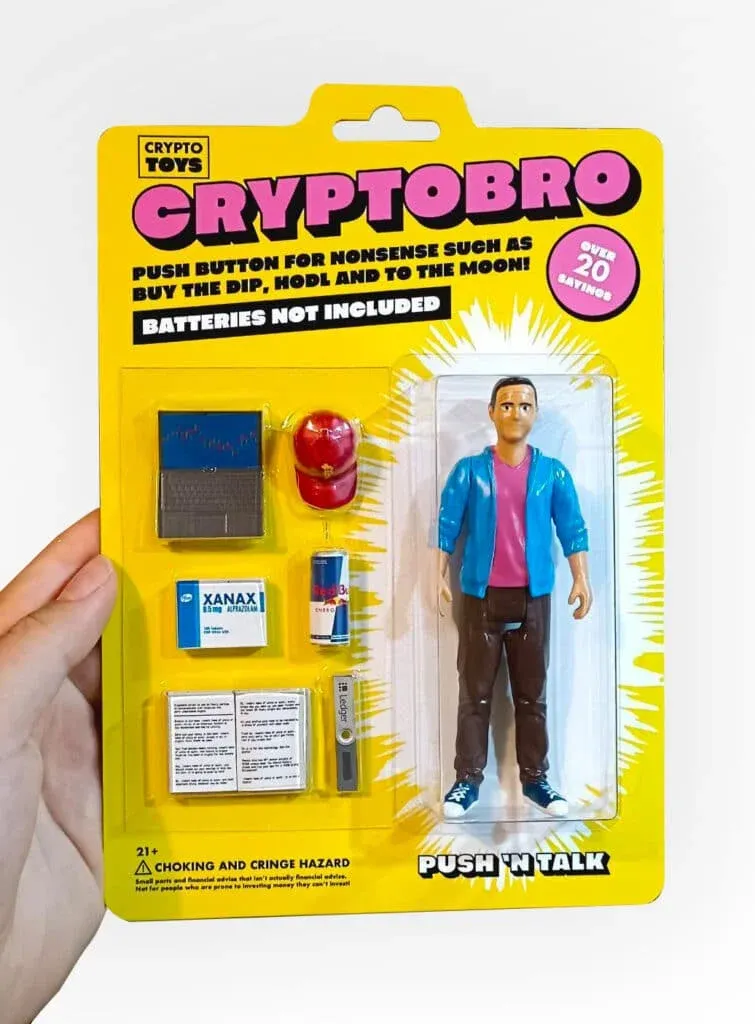 A novelty toy package labeled 'CRYPTObro' containing a figurine dressed in casual clothes, along with accessories like a laptop, a cap, a can of energy drink, a box labeled 'Xanax,' a book, and a USB device. The packaging humorously mimics collectable action figures, with text like 'Push button for nonsense such as buy the dip, hodl and to the moon!' and 'Batteries not included.' There's also a warning about 'choking and cringe hazard.' The design is colorful with bold yellow and pink accents.
