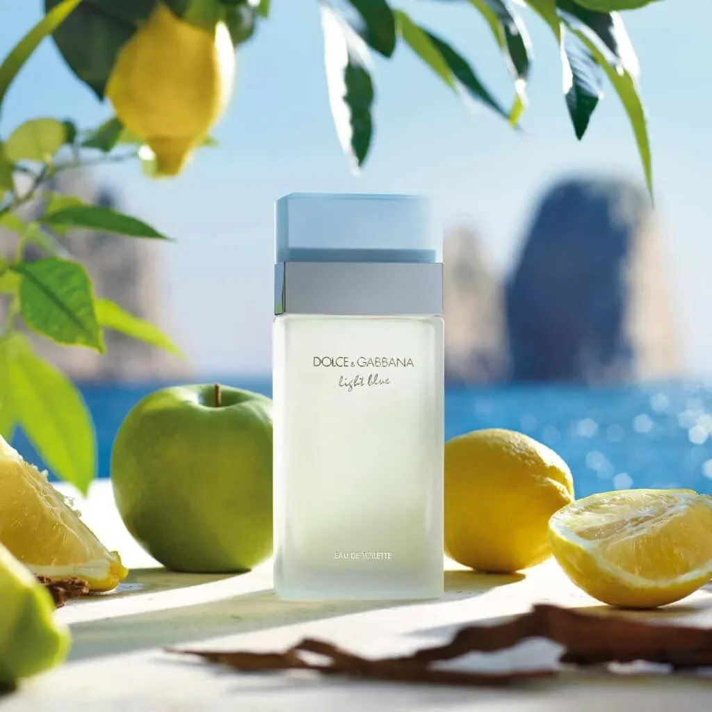 A bottle of Dolce & Gabbana Light Blue perfume is placed on a surface with lemons and a green apple, set against a scenic ocean view with clear skies and distant rock formations.