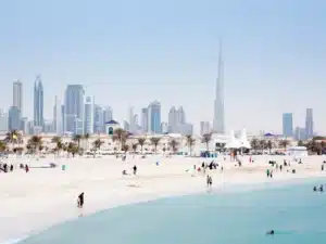 A beach with people enjoying the sand and water; in the background is a city skyline with modern skyscrapers and a tall tower, under a clear blue sky.