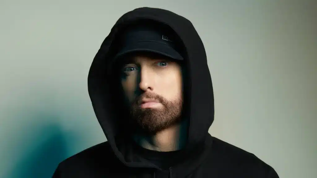A person wearing a black hoodie and a black cap, with a neutral expression, looking towards the camera.