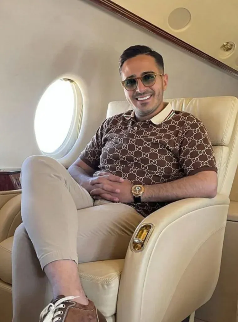 A person sitting comfortably in a luxury airplane seat, wearing sunglasses, a patterned shirt, beige pants, and brown shoes, smiling towards the camera.
