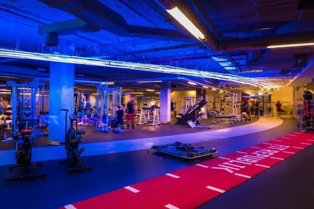 A large, modern gym with various exercise machines and equipment, illuminated by blue ambient lighting. A red exercise mat is on the floor in the foreground. Several people are engaged in workouts on different equipment.