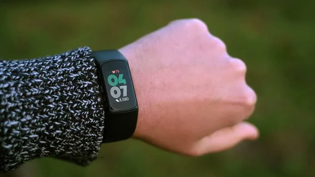 A left hand wearing a black fitness tracker displaying the time as 4:07, a heart rate of 79 bpm, and a step count of 8,580; the person is wearing a knitted sweater with a black and white pattern.