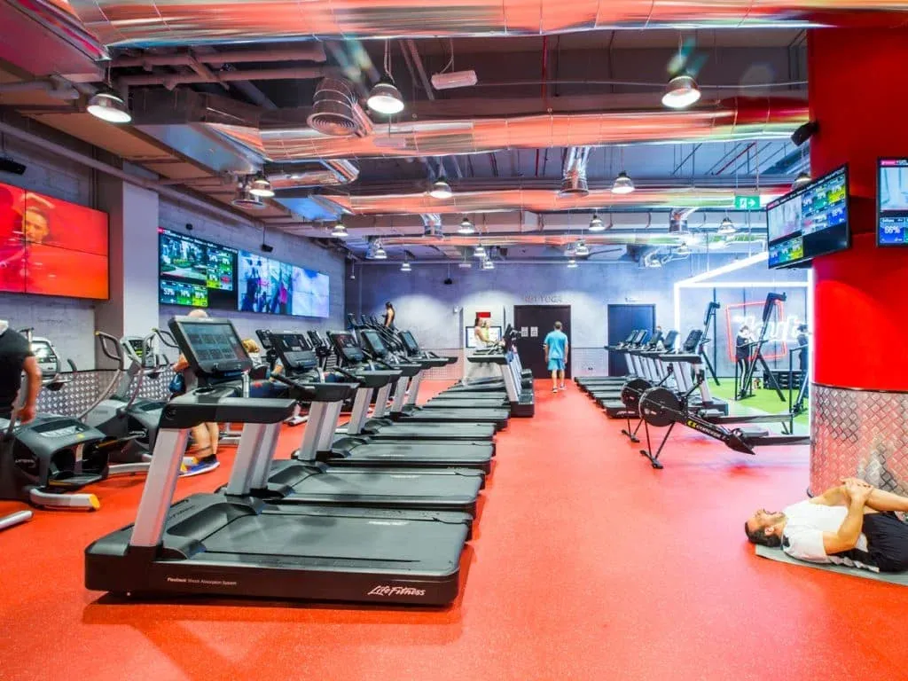 A modern gym interior with multiple treadmills lined up on a red floor, wall-mounted screens displaying various channels, and a man stretching on a mat in the foreground.