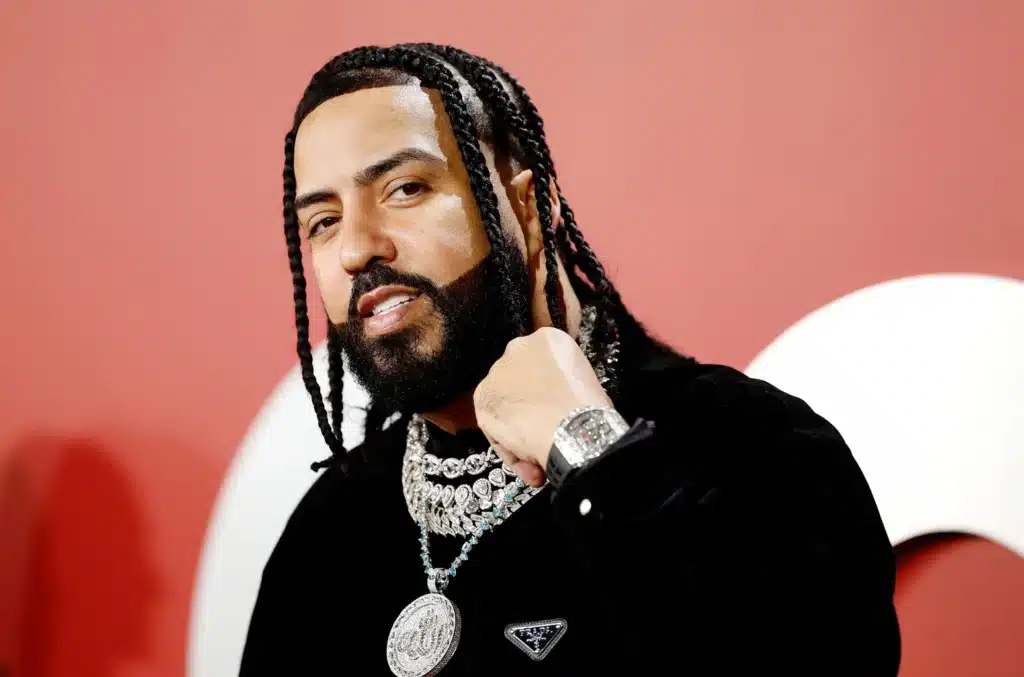 Person with braided hair and a beard, wearing a black outfit, several necklaces, and a watch, standing against a red background.