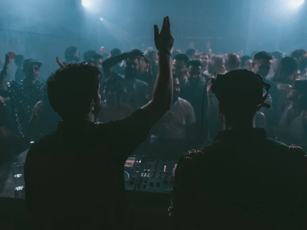 Two DJs performing in a dimly lit club with a crowded dance floor and raised hands in the audience.