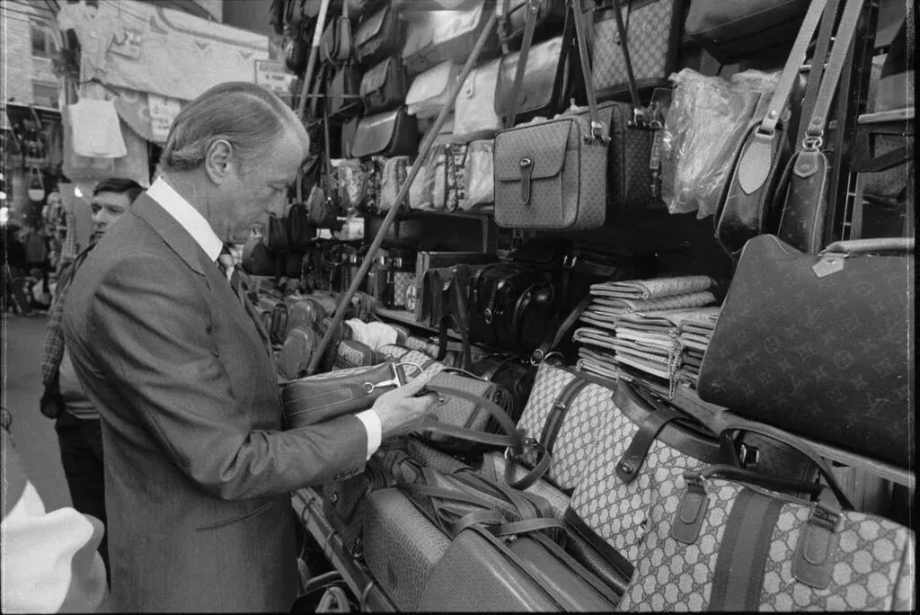 A man in a suit examines handbags at a market stall displaying various luxury bags and accessories.