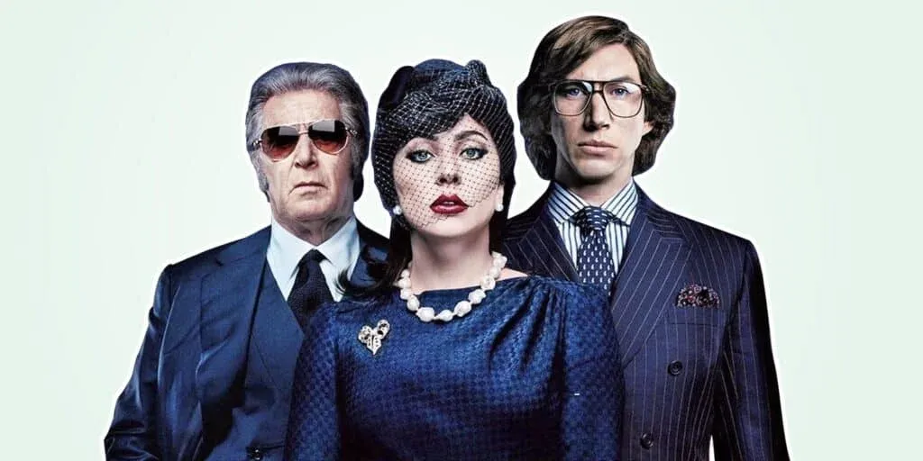 Three people in formal attire pose for a photo; the person on the left wears sunglasses and a dark suit, the person in the center wears a blue dress with a hat and pearl necklace, and the person on the right wears glasses and a pinstripe suit.