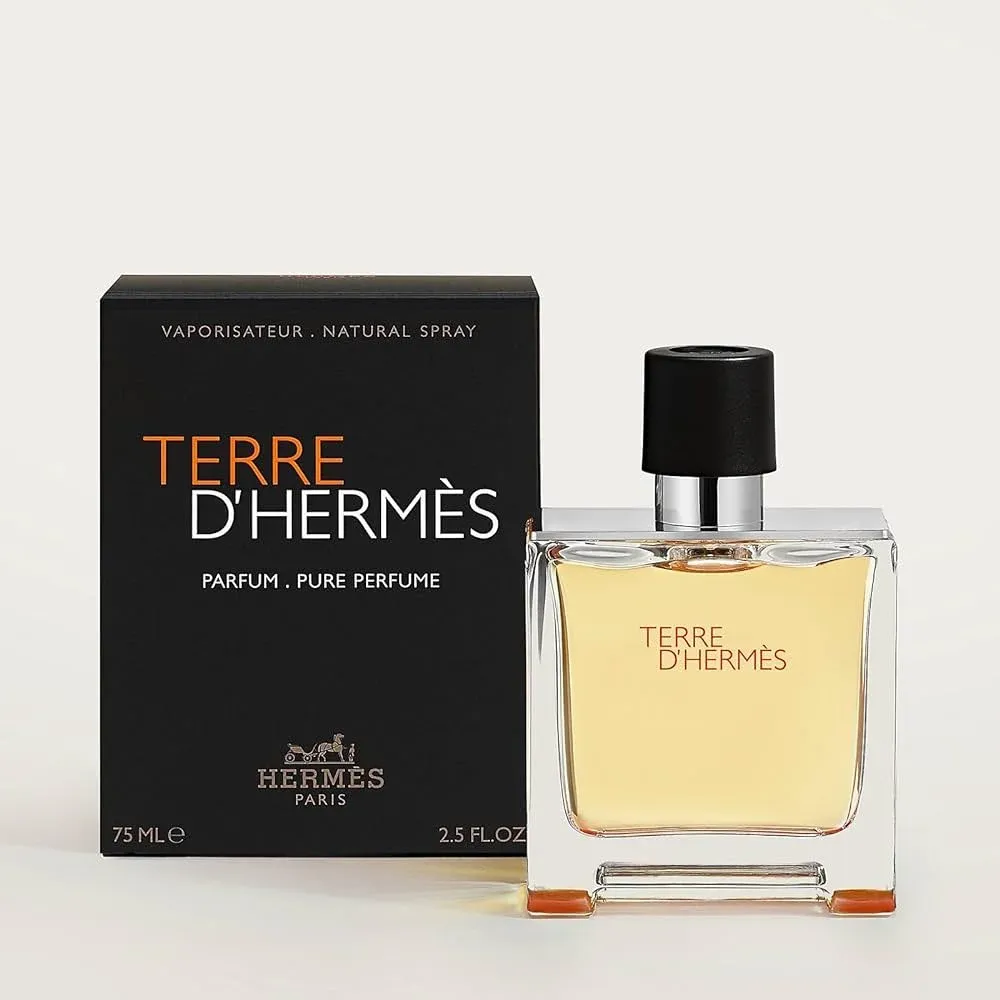 A bottle of Terre d'Hermès perfume standing in front of its black packaging. The perfume bottle is clear with a black cap, and the text on both bottle and packaging is in contrasting orange and white colors.