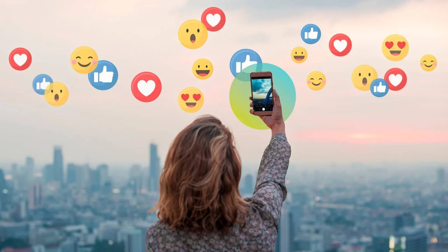 A person takes a selfie with a smartphone against a city skyline at sunset, surrounded by floating social media reaction icons such as likes, hearts, and smiley faces.