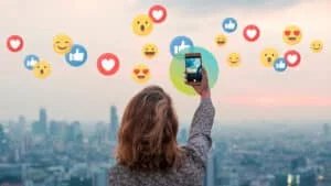 A person takes a selfie with a smartphone against a city skyline at sunset, surrounded by floating social media reaction icons such as likes, hearts, and smiley faces.