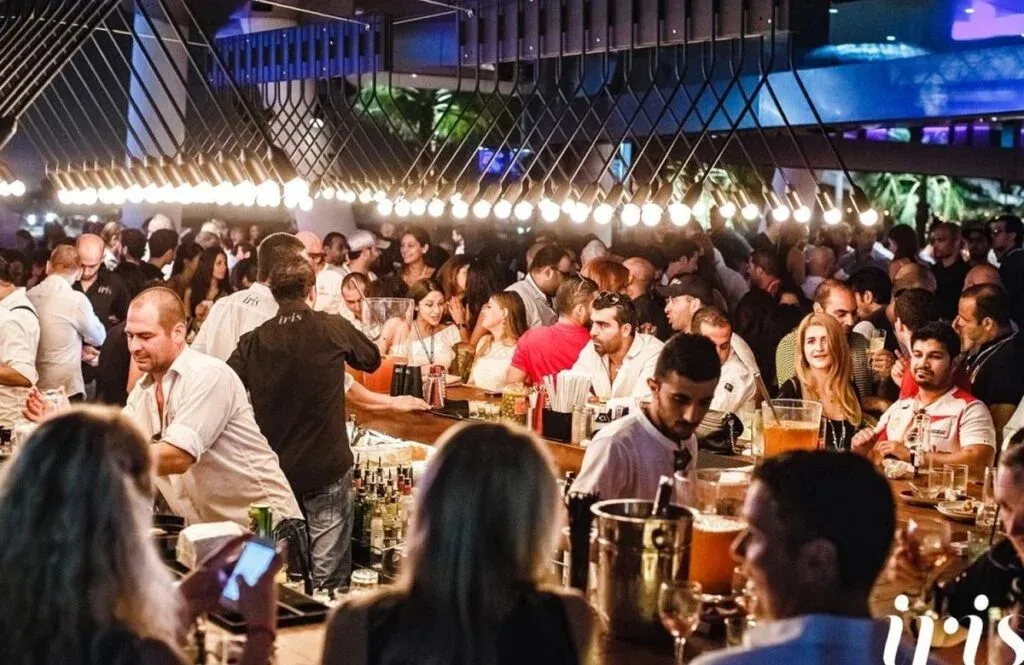 A crowded bar scene with people socializing and bartenders serving drinks under modern pendant lighting. The atmosphere is lively and bustling.