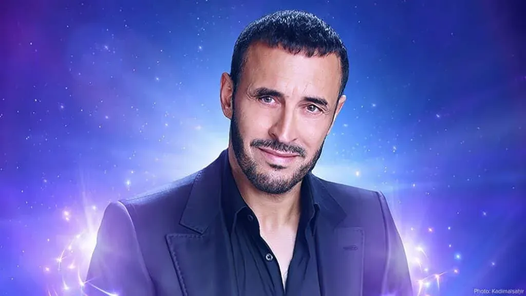 A man with short dark hair and a beard smiling, wearing a dark suit against a cosmic, starry background.