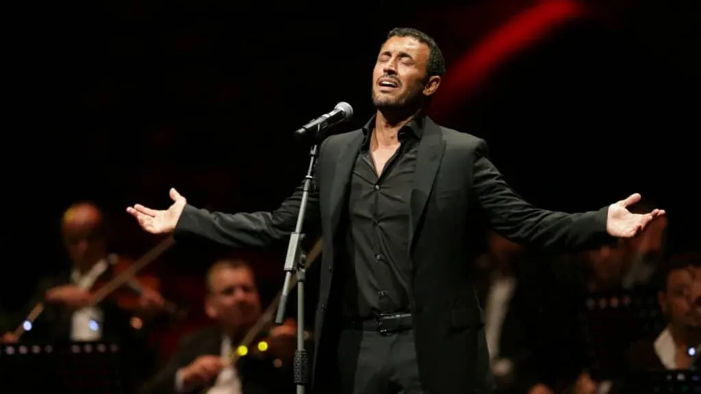 A man in a black suit passionately singing into a microphone on stage, with an orchestra in the background.