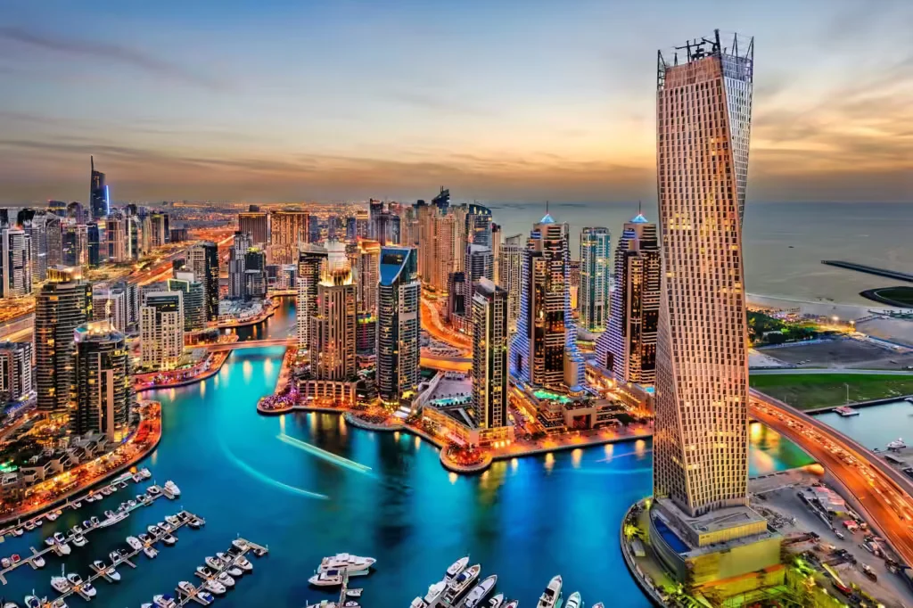 Aerial view of a vibrant city skyline with a prominent twisted skyscraper near the waterfront, surrounded by numerous high-rise buildings and illuminated at twilight, with a marina filled with boats in the foreground.