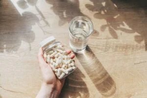 A hand holding a clear jar filled with white capsules next to a glass of water on a wooden surface, with leaf shadows cast on the table.