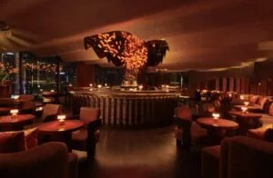A dimly lit, upscale lounge with plush seating and a circular bar at the center, featuring warm ambient lighting and a decorative ceiling fixture above the bar.