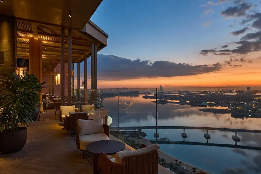 A luxurious outdoor terrace with cushioned seating, overlooking a cityscape and calm water at sunset. The sky is painted in warm hues of orange and blue, with modern architecture visible in the distance.
