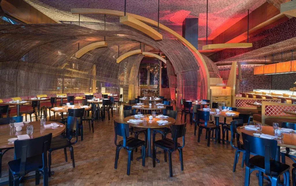 A modern restaurant interior with round wooden tables and black chairs, featuring an intricate ceiling design and a unique arching structure made of woven materials and wooden beams, with soft red lighting creating a warm ambiance.