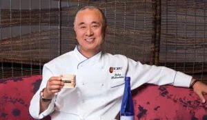 A chef in a white coat with the logo 'Nobu Las Vegas' holding a small wooden box, seated on a red-patterned couch with a decorative woven backdrop.