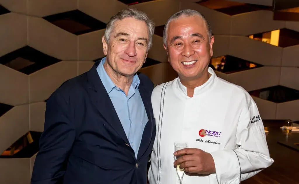 Robert De Niro and Nobu Matsuhisa standing close together in a warmly lit room; the man on the left is wearing a dark suit with a blue shirt, and the man on the right is wearing a white chef's jacket with "Nobu Caesars Palace" embroidered on it, holding a glass of champagne.