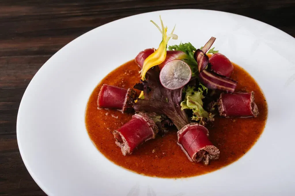 A gourmet dish featuring rolled slices of cured meat arranged around a pile of mixed greens, garnished with carrot ribbons and a thin slice of radish, served on a bed of rich, brown sauce.