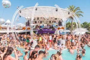 A lively pool party with a large crowd of people enjoying the sun, swimming, and dancing around a DJ stage with a "Kisstory" sign. The scene includes decorative structures, palm trees, and a vibrant atmosphere under a clear blue sky.