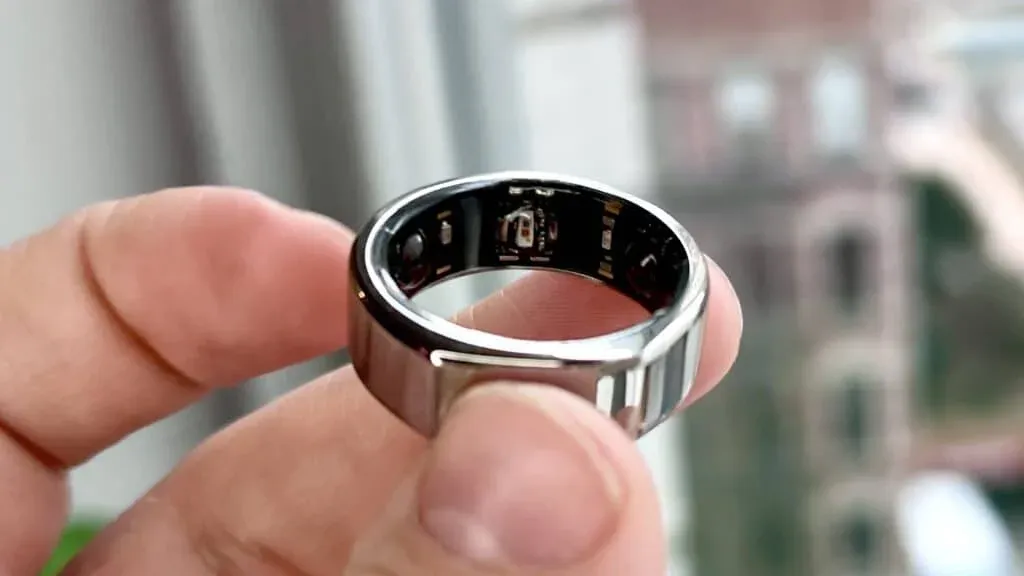 A close-up view of a person's hand holding a metallic smart ring, showing the inner components and sensors inside the band, against a blurred background.