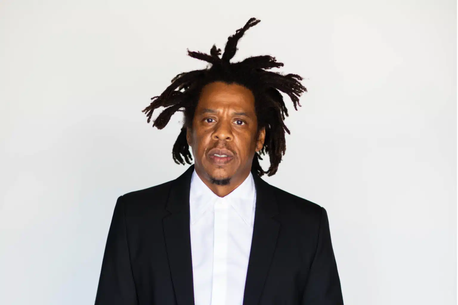A man wearing a black suit and white shirt stands against a plain white background, with distinctive, textured hair styled upwards.