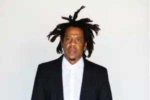 A man wearing a black suit and white shirt stands against a plain white background, with distinctive, textured hair styled upwards.