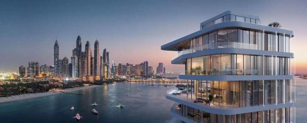 Modern multi-story building with large glass windows overlooking a calm waterfront at sunset, with a city skyline in the background.