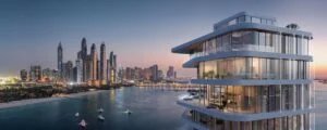 Modern multi-story building with large glass windows overlooking a calm waterfront at sunset, with a city skyline in the background.