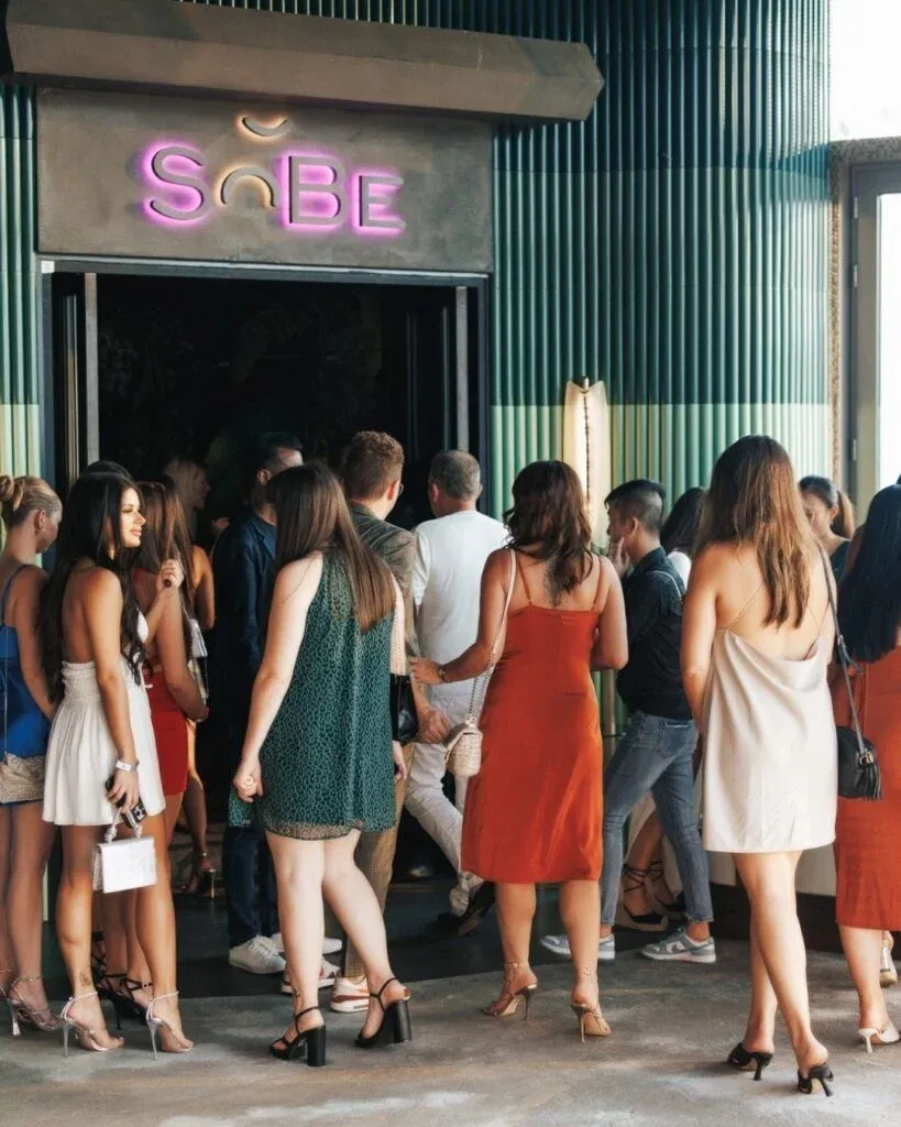 A group of people, mostly women in dresses and high heels, are gathered outside a building with a sign reading 'SoBe' in pink neon letters.